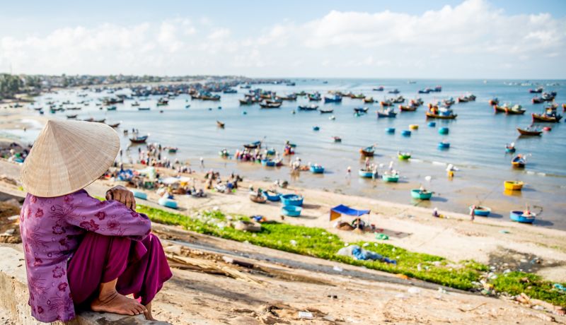 femme au chapeau conique assise sur la plage animée de Mui Ne